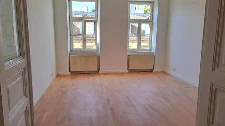 Helles, leeres Zimmer mit Parkettboden und zwei großen Fenstern, die auf die Straße blicken.