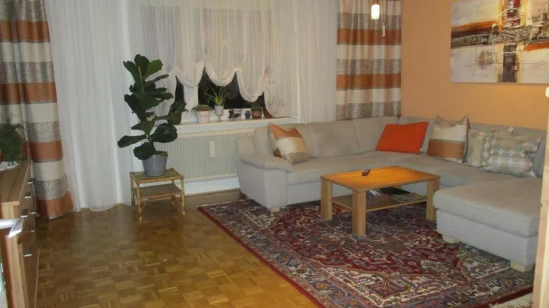 Gemütliches Wohnzimmer mit Parkettboden, beiger Eckcouch und gestreiften Vorhängen.