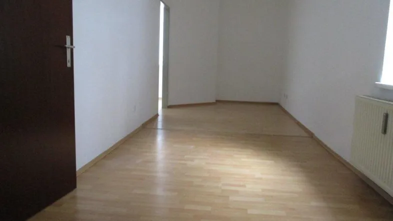 Helles, leeres Zimmer mit hellem Laminatboden, weißen Wänden und einem Heizkörper unter dem Fenster.