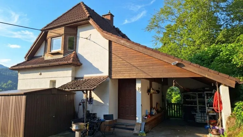Vorderansicht des Hauses mit Carport, Schuppen und Satteldach unter blauem Himmel.