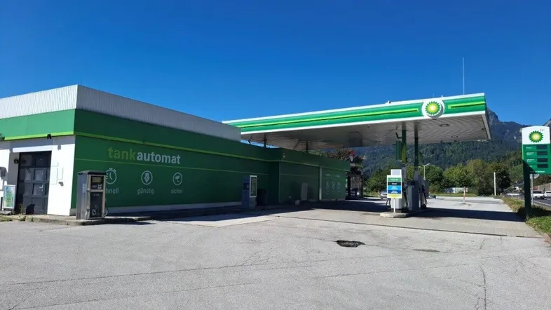 Tankstelle mit grünem Gebäude und Tankautomat, umgeben von Bergen unter blauem Himmel.