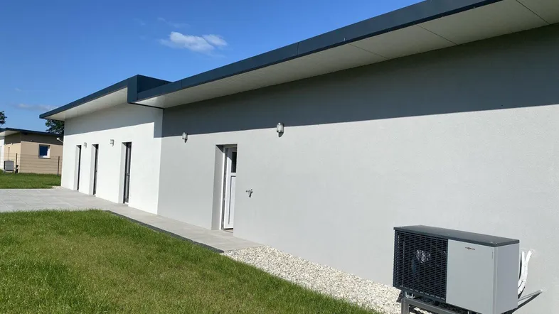 Moderne Hausfassade mit Flachdach, weißem und grauem Putz sowie einer außenliegenden Wärmepumpe.