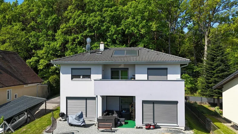 Modernes Einfamilienhaus mit Satteldach, Balkon und Terrasse, umgeben von viel Grün und Bäumen.