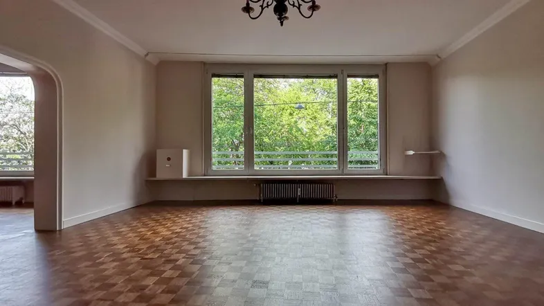 Helles Wohnzimmer mit Parkettboden, Stuckdecke und Blick ins Grüne durch große Fenster.