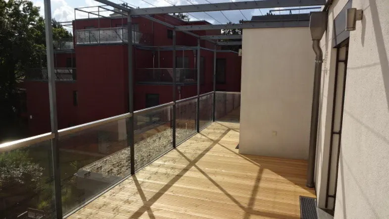 Große Holzterrasse mit Glasgeländer und Blick auf umliegende Gebäude unter blauem Himmel.