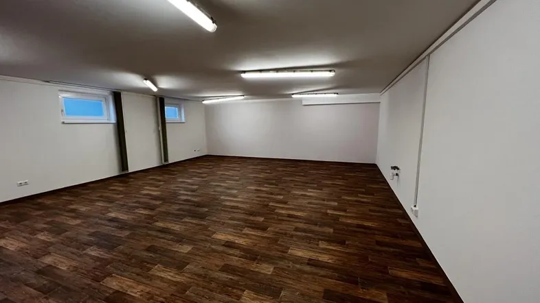 Geräumiger, leerer Raum mit dunklem Holzdekorboden, weißen Wänden und kleinen Fenstern, beleuchtet von Leuchtstoffröhren.