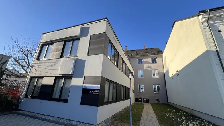 Modernes Bürogebäude mit grauer Fassade, großen Fenstern und gepflastertem Zugangsbereich unter blauem Himmel.