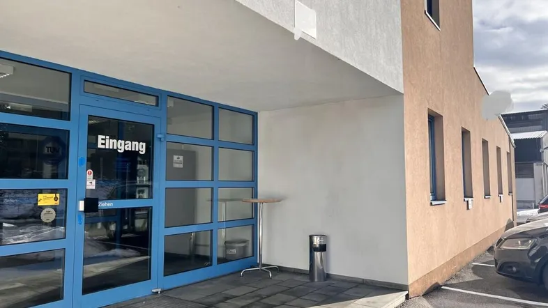 Eingangsbereich eines modernen Geschäftsgebäudes mit blauen Glastüren und einem kleinen Stehtisch davor.