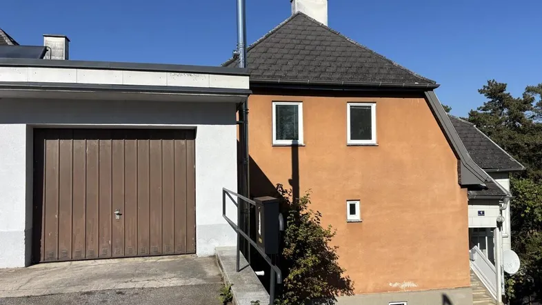 Seitenansicht eines Hauses mit angeschlossener Garage und schräger Zufahrt unter blauem Himmel.