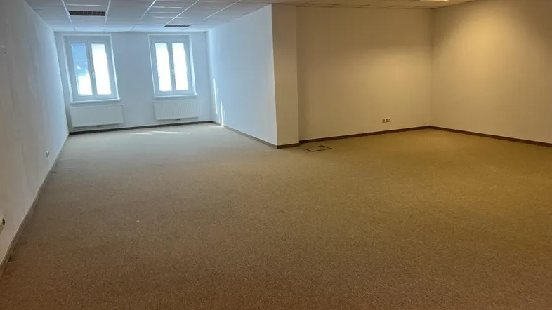 Geräumiger, leerer Büroraum mit Teppichboden und großen Fenstern, die viel Tageslicht hereinlassen.