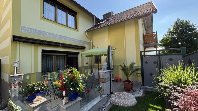 Sonnige Terrasse mit Gartenmöbeln, Sonnenschirm und Blumen vor dem Haus.