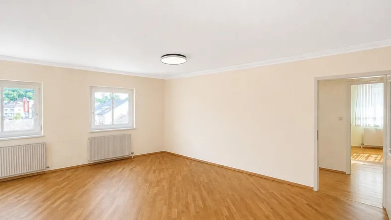 Helles Zimmer mit Parkettboden, beigen Wänden und Blick ins Grüne durch die Fenster.
