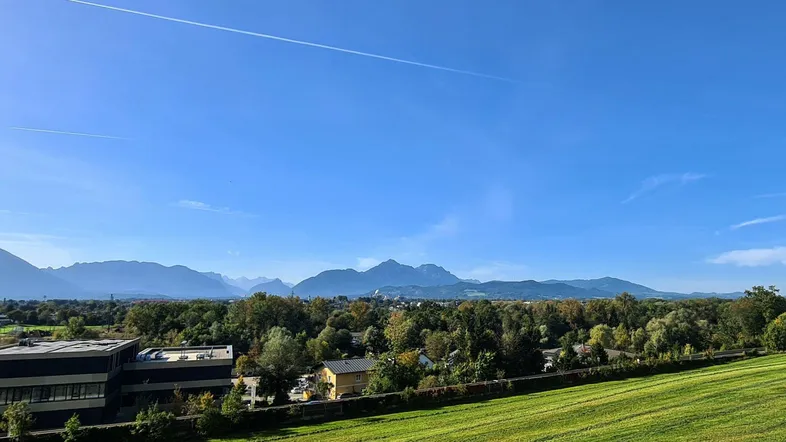 Panoramablick über eine weite grüne Wiese, Baumreihen und eine Stadt mit Bergen im Hintergrund unter klarem Himmel.