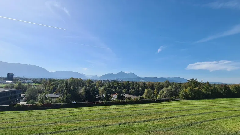 Weitläufiger Blick über grüne Felder, Gebäude und eine beeindruckende Berglandschaft unter blauem Himmel.