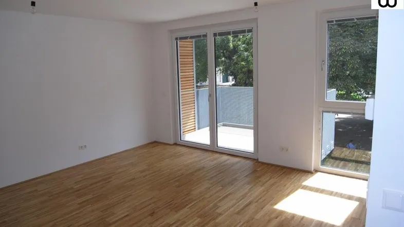 Helles Zimmer mit Holzboden, weißen Wänden und Zugang zu einem Balkon.