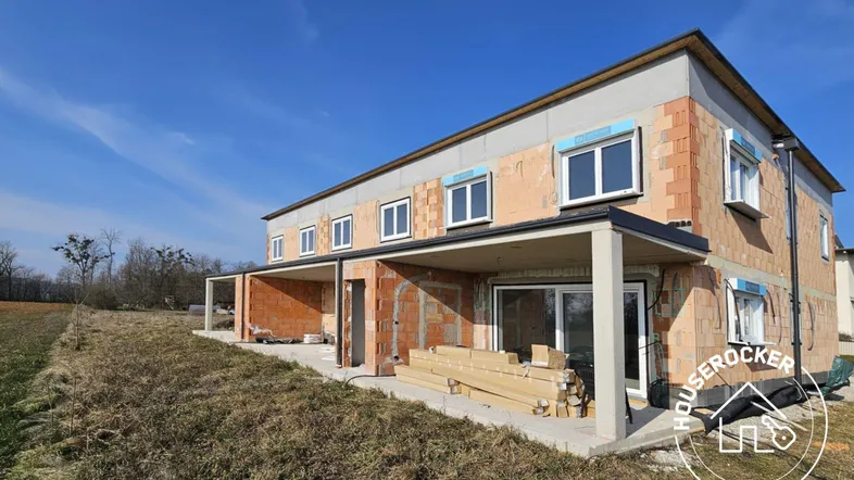 Rohbau eines modernen Reihenhauses mit großen Fensteröffnungen und zukünftigen Terrassenbereichen unter blauem Himmel.