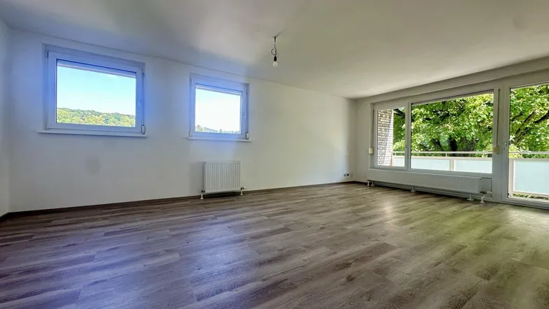 Helles, leeres Zimmer mit Laminatboden und Zugang zum Balkon, umgeben von grünen Bäumen.