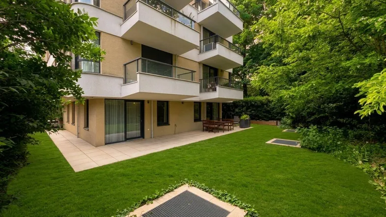 Modernes Gebäude mit gepflegtem Rasen, großer Terrasse und Balkonen, umgeben von viel Grün.