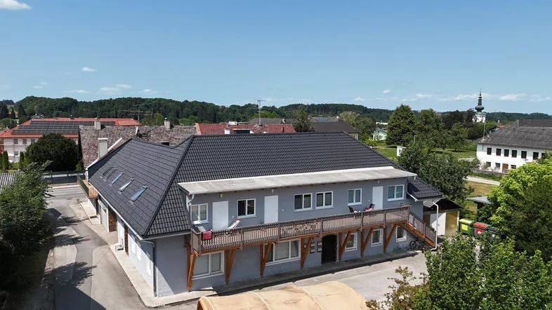 Luftaufnahme eines Gebäudes mit dunklem Dach, heller Fassade und langem Holzbalkon in ländlicher Umgebung.