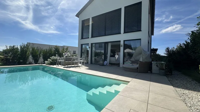 Modernes Haus mit privatem Pool und Terrasse, umgeben von grüner Landschaft unter blauem Himmel.