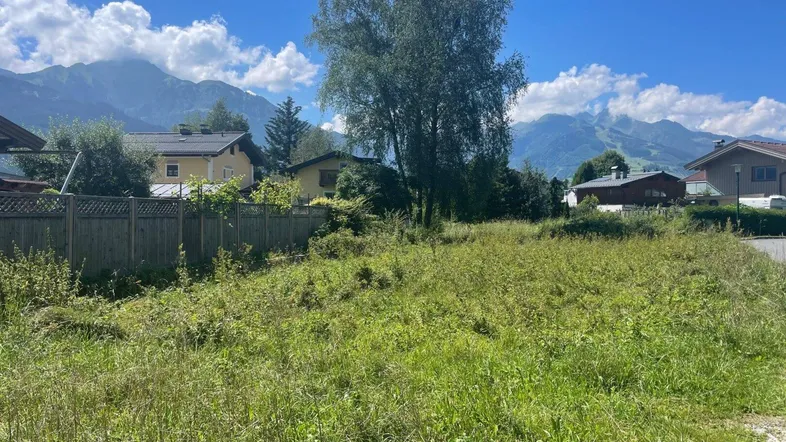 Grünes Grundstück mit Bergblick und umliegenden Häusern unter blauem Himmel an einem sonnigen Tag.