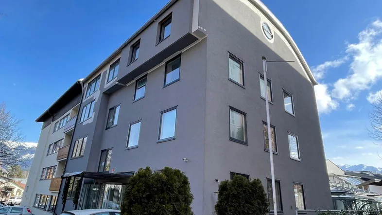 Modernes Mehrfamilienhaus mit geschwungener Dachlinie und grauer Fassade, umgeben von Bergen.