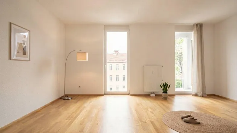 Heller, leerer Wohnraum mit großen Fenstern, Holzboden und viel Tageslicht.