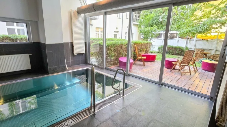 Innenpoolbereich mit großen Glasfronten und direktem Zugang zu einer möblierten Außenterrasse.