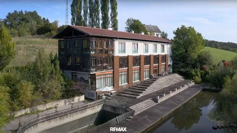 Großes Gebäude mit einzigartiger Fassade und Holzdeck am Wasser, umgeben von grüner Landschaft.