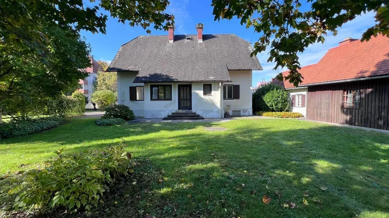 Traditionelles Haus mit großem Vorgarten und altem Baumbestand unter blauem Himmel.