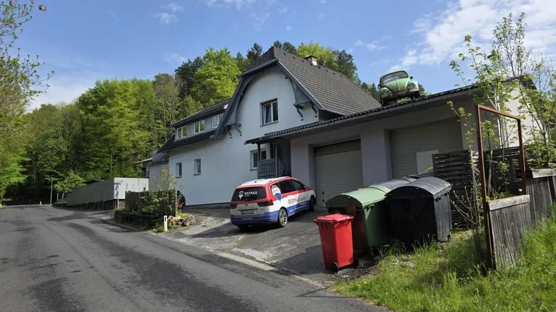 Ansicht des Hauses von der Straße mit Garagen, einem geparkten Auto und Mülltonnen.