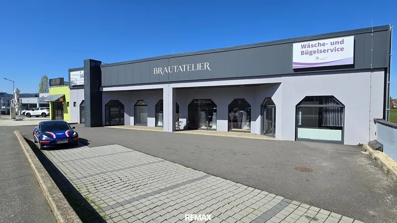 Modernes Geschäftsgebäude mit großen Schaufenstern und einem gepflasterten Parkplatz davor unter blauem Himmel.