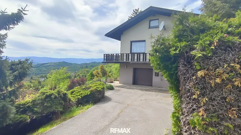 Hausfassade mit Balkon und Auffahrt, umgeben von üppiger Natur und weitem Ausblick.