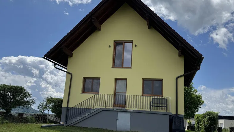 Seitenansicht des gelben Hauses mit rotem Dach, Außentreppe und einem kleinen Balkon unter blauem Himmel.