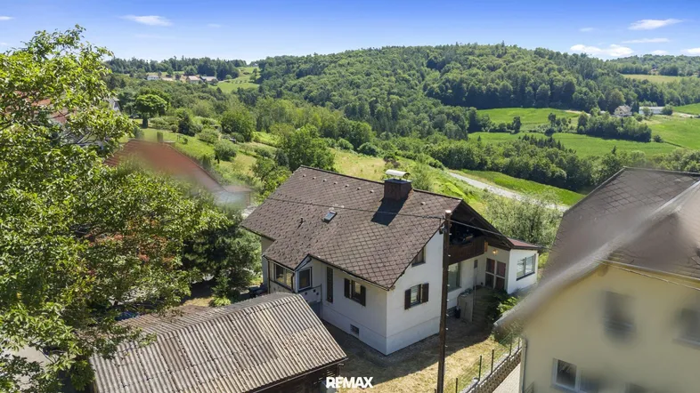 Luftaufnahme eines Hauses in einer grünen, hügeligen Landschaft mit umliegenden Wäldern.
