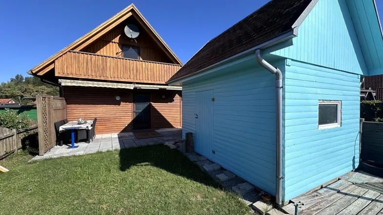 Außenansicht eines Holzhauses mit angrenzendem blauen Schuppen, gepflasterter Terrasse und Rasenfläche.