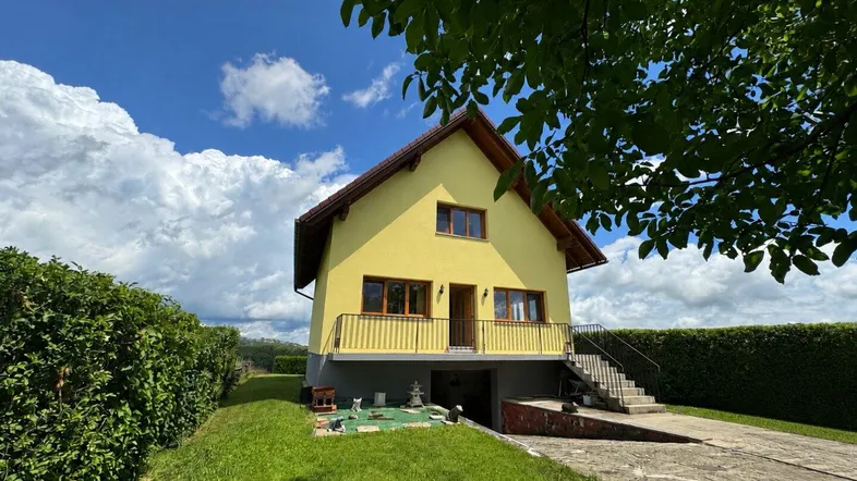 Gelbes Einfamilienhaus mit Satteldach, Balkon und großem Garten unter blauem Himmel an einem sonnigen Tag.