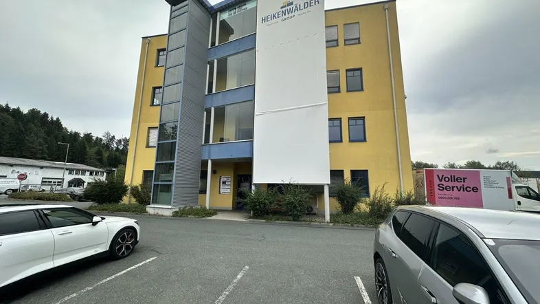 Gelbes Bürogebäude mit moderner Glasfassade und Außenparkplätzen unter bewölktem Himmel.