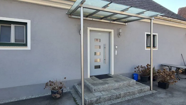 Eingangsbereich eines Hauses mit modernem Vordach aus Glas und Metall über Steinstufen.