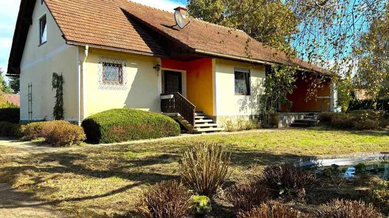 Gelbes Einfamilienhaus mit braunem Ziegeldach, umgeben von einem gepflegten Garten mit Teich.