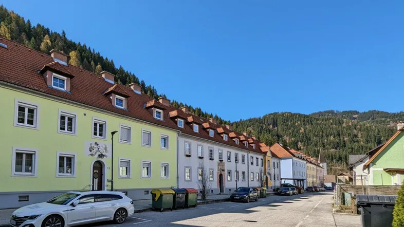 Eine ruhige Wohnstraße mit mehrstöckigen Gebäuden und bewaldeten Hügeln im Hintergrund unter blauem Himmel.