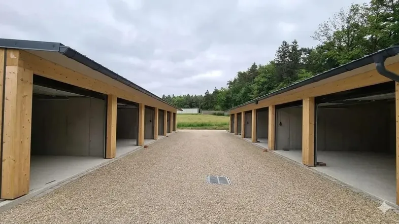Zwei Reihen neuer Garagenboxen mit Holzbalken und Schotterzufahrt, umgeben von grüner Landschaft.