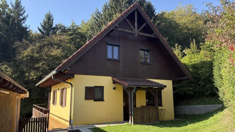 Hausfassade in Gelb mit Holzelementen und Satteldach, umgeben von viel Grün und Bäumen.
