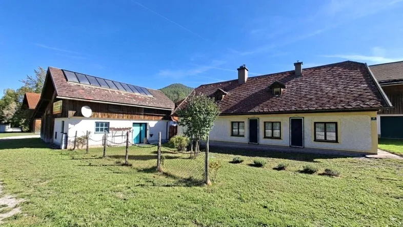 Traditionelle Gebäude mit Satteldächern und Solaranlage, umgeben von grünen Wiesen und Bergen unter blauem Himmel.