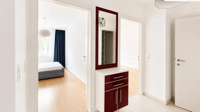 Heller Flur mit Holzspiegel und Blick in ein Schlafzimmer mit Holzboden.