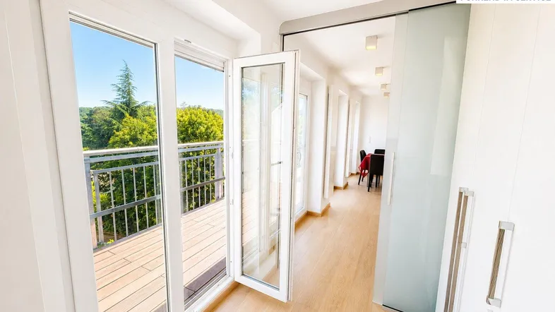 Heller Flur mit Holzboden und großen Fenstern, die Zugang zu einem Balkon bieten.