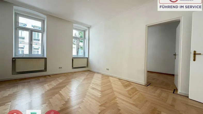 Helles Zimmer mit Fischgrätparkettboden und zwei großen Fenstern, das als Wohnzimmer genutzt werden könnte.