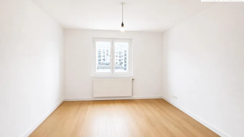 Helles, leeres Zimmer mit Parkettboden und einem Fenster, das Tageslicht hereinlässt.