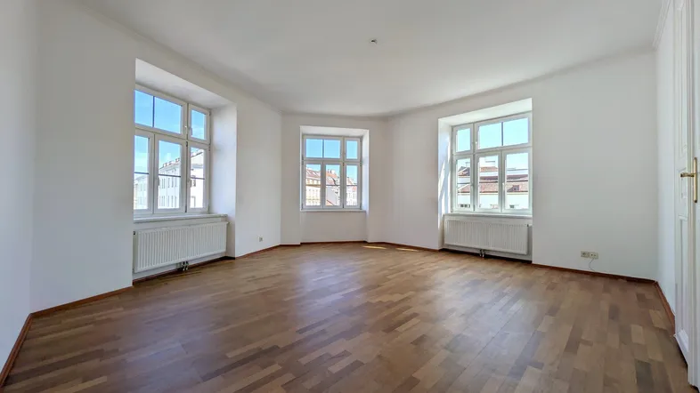 Helles Zimmer mit Fischgrätparkett, weißen Wänden und großen Fenstern, die viel Tageslicht hereinlassen.