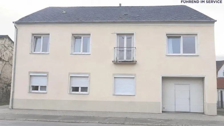Beigefarbene Fassade eines Mehrfamilienhauses mit Satteldach, kleinem Balkon und Garagentor.